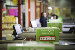 A row of checkout counters in a store displaying a sign that reads "Lipa na M-Pesa Accepted Here" with a till number "893225" prominently shown. In the background, there are blurred figures of people and other checkout stations.